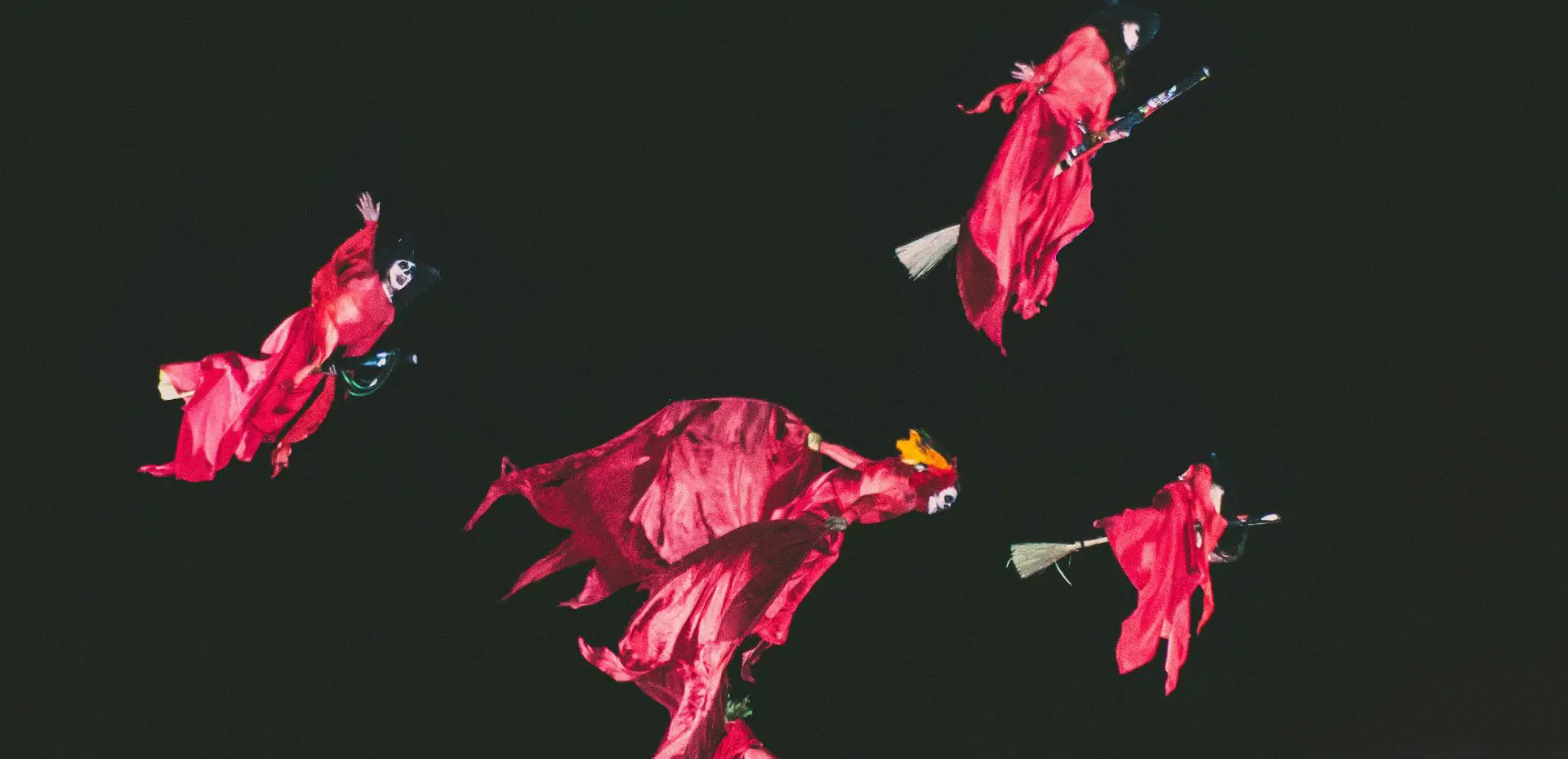 Quattro figure in abiti rossi da strega volano nella notte su scope di legno, sospese in un cielo nero: una scena teatrale, queer e ironica che evoca magia lesbica, gioco collettivo e libertà ribelle.