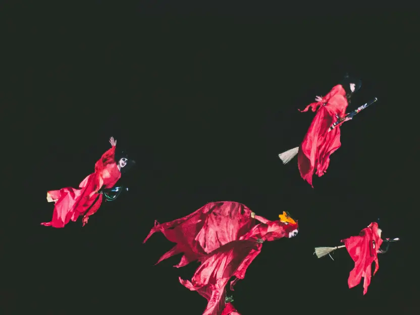 Quattro figure in abiti rossi da strega volano nella notte su scope di legno, sospese in un cielo nero: una scena teatrale, queer e ironica che evoca magia lesbica, gioco collettivo e libertà ribelle.