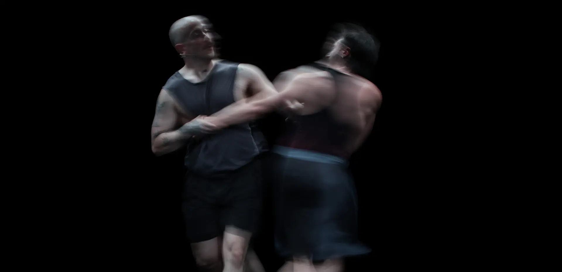 Due danzatori in abiti scuri si afferrano in un abbraccio dinamico che preannuncia l'inizio di una vorticosa danza in cerchio, il palco è buio, l'immagine sfocata, l'atmosfera intensa e fluida che trasmette forza corporea, tensione emotiva e dinamismo.