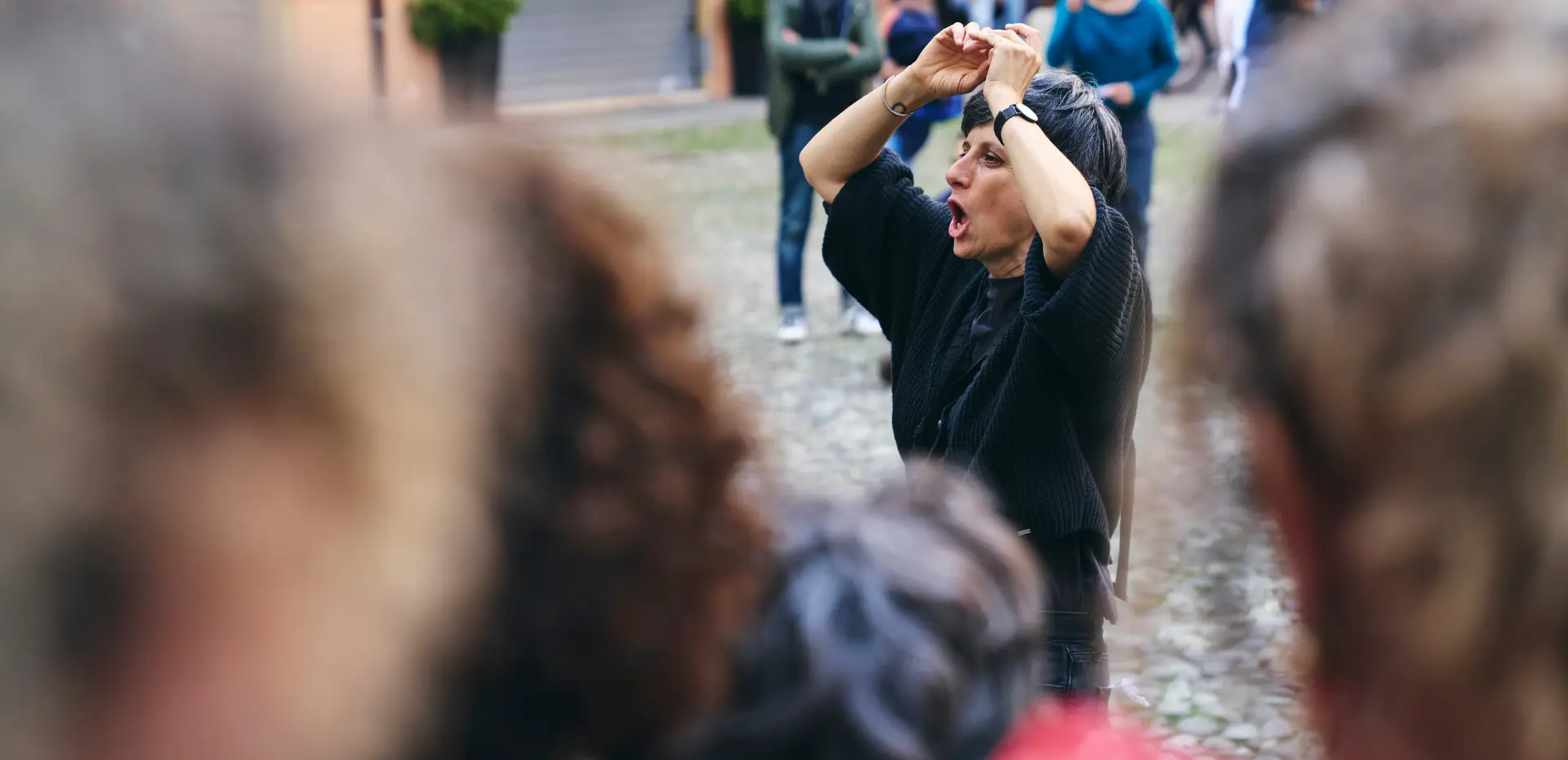 Persona che alza le braccia in gesto espressivo davanti a un piccolo gruppo in strada, tra teste sfocate in primo piano: un laboratorio urbano, intimo e partecipato, che invita all’ascolto del corpo e dell’altro.