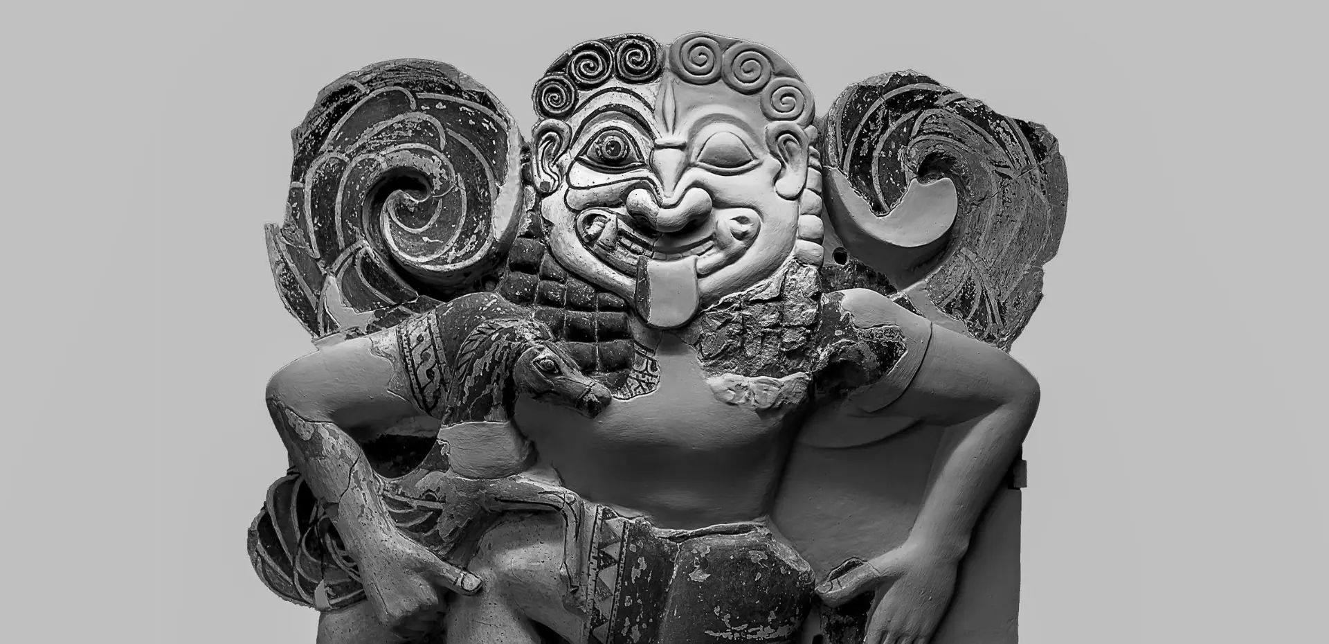 Fotografia in bianco e nero di un'antica scultura in terracotta raffigurante una figura simile a una gorgone, con un viso ghignante, lingua sporgente e grandi elementi decorativi a spirale ai lati, mentre tiene degli animali tra le braccia.