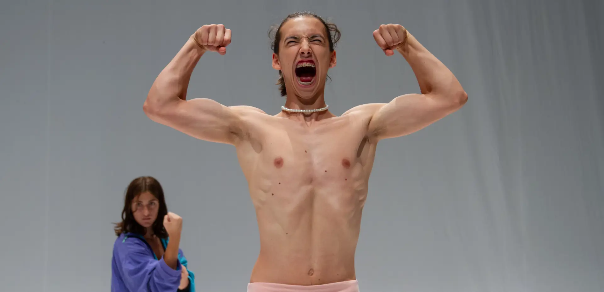 Persona a torso nudo in primo piano posa con le braccia flesse in segno di forza e la bocca spalancata come in un grido. Indossa una collana di perle e un indumento rosa con la scritta “Unicorn.”. Sullo sfondo, leggermente sfocata, un’altra persona con una giacca viola tiene un pugno alzato in una posa combattiva. Lo sfondo è uniforme e chiaro.