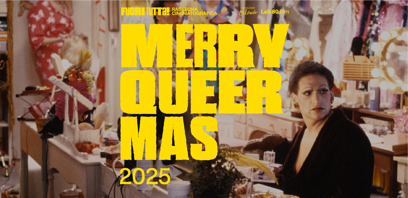 Interno di un camerino affollato e colorato: tavoli da trucco pieni di parrucche, trucchi, fiori e oggetti scenici. In primo piano una persona seduta davanti allo specchio illuminato guarda verso sinistra con un’espressione seria. Sullo sfondo si vedono altre persone e costumi variopinti appesi alle pareti, creando un’atmosfera vivace tipica del backstage di uno spettacolo drag